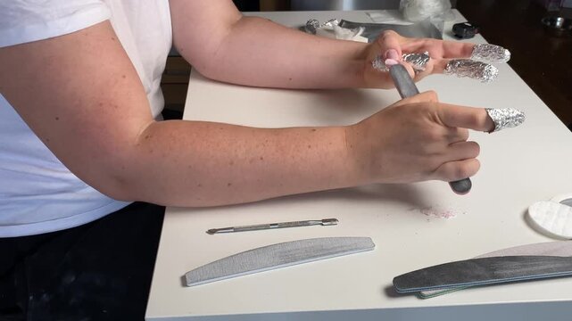 filing nails and removing gel polish with foil wraps on fingertips. woman performing manicure at home using nail file and remover tools. concept of nail care, beauty routine, diy manicure