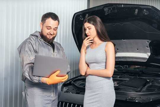 Car mechanic communicating with a female customer while using laptop and examining vehicle breakdown at auto repair shop