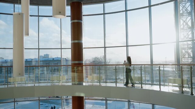 Wide view of stylish modern mall interior with large glass panels and natural daylight as elegant woman walks gracefully across upper floor walkway balancing book on head with confident posture
