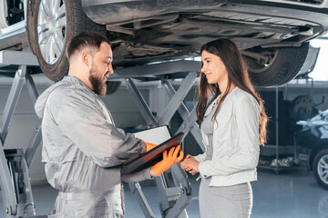 Auto Mechanic holding clipboard talking to female customer in workshop at auto repair center. Quality of Service