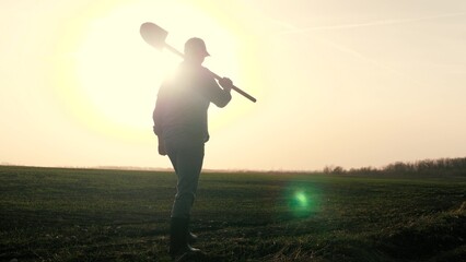 An agronomist man walks through a black plowed field at sunset, farmer with a shovel in his hands, work in the field for the worker, dawn time in the sky for life activities in agriculture. © TO LOVE
