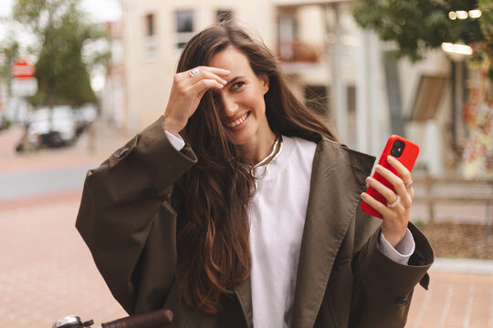 Brunette woman standing near bicycle and hold phone in city, street or urban road outdoors. Bike, travel and happy female using 5g mobile tech, internet browsing or social media, fix her hair.