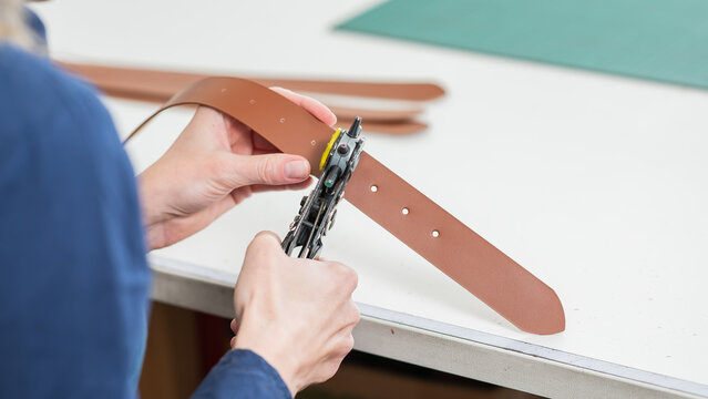 A woman makes holes in a leather belt. Leatherworker's workshop. 