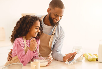 Best Recipe App. Smiling young bearded African American father embracing his little daughter and using digital tablet in kitchen, choosing what to cook, showing website, curly girl mixing ingredients