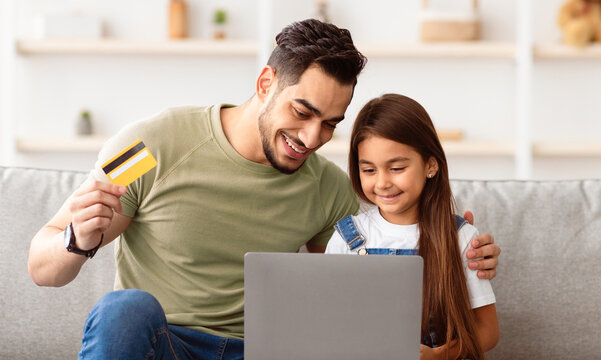 Online Shopping Concept. Portrait of cheerful daddy and daughter using computer, holding and showing debit credit card, sitting on sofa in living room making purchase on Internet, looking at screen