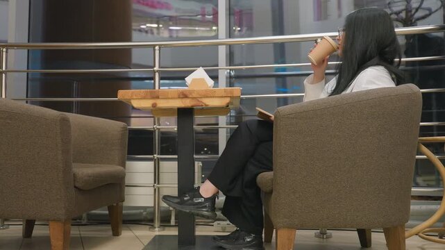 Investor seated in restaurant picks up drink, takes sip, returns cup to table and resumes reading book, sitting comfortably in armchair with relaxed posture, with evening city lights visible