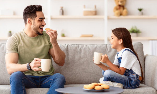 Snack Time. Portrait of cheerful little girl and man holding cups, drinking coffee and tea, eating cookies and looking at each other, dad and daughter talking sitting on the couch in living room
