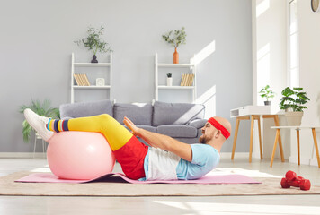 Funny young man in sportswear having home workout lying on floor on yoga mat with fit ball. Sporty...