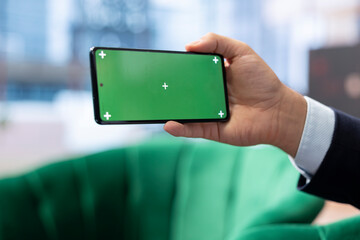 Determined male administrator uses phone with copy space display, tracking profit margins and making data driven decisions. Young entrepreneur working on a success story, multitasking.