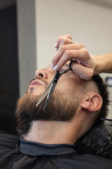 Barber shaving bearded customer in modern style barbershop. Male Beard hair cutting with scissors close up. Man client visit gentleman grooming beauty salon for evens the beard