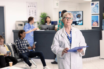 Fototapeta premium Elderly doctor stands in hospital lobby with clipboard in hand, prepared to consult with various patients. Caucasian female physician wearing lab coat and looking directly at camera.