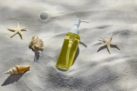 A clear skincare product bottle sits on white sand among seashells