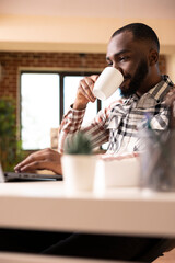 Black man entrepreneur drinking coffee and reading online articles on laptop. African american male copywriter proofreads marketing material and analyzes business data from home office.