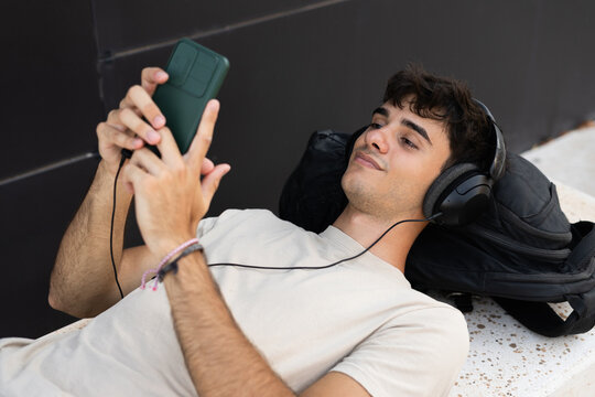 Young man using cell phone and listening to music on a bench outdoors