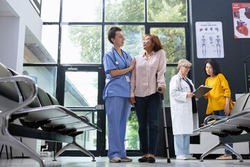 Obraz premium Nurse supporting asian patient while she walks in crutches during physical therapy in hospital waiting area. Medical expert helping woman with locomotor issues, healing process.