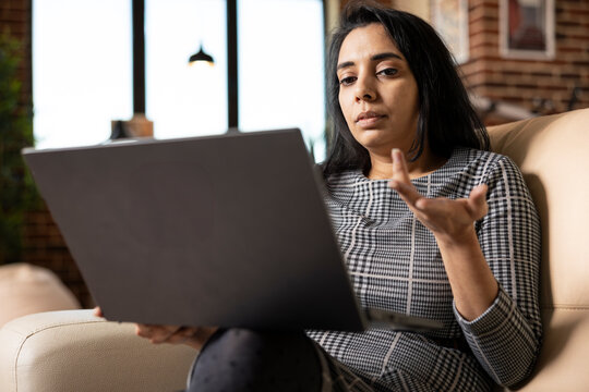 Female consultant working from home, seated on sofa discussing business projections with client on video call. Indian woman entrepreneur sharing marketing ideas during virtual meeting with colleagues.
