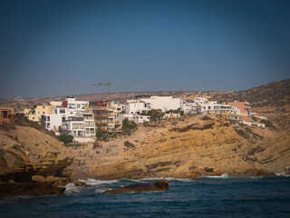 Fototapeta premium Village with modern, white buildings built along rocky, sandy cliffside. Scenic coastal view of holiday destination of Imsouane, Morocco where gentle waves of Atlantic Ocean roll onto the shore below