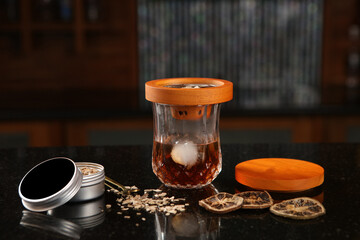 A sophisticated drink is shown in a glass with a wooden smoking top and an ice cube. The scene includes dried citrus slices and wood chips, highlighting a craft cocktail experience