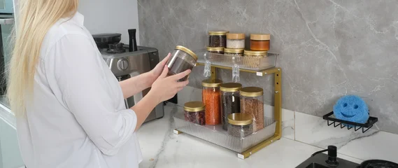 A woman in a white shirt organizes her kitchen counter, placing a spice jar onto a stylish two-tiered golden rack. The image showcases smart kitchen storage and home organization. © Imageologist