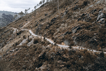 Narrow winding dirt road cutting through steep rocky mountain slope covered with dry shrubs, sparse trees, and rugged stone formations under cloudy sky; autumn Camino de Santiago in Asturias, Spain