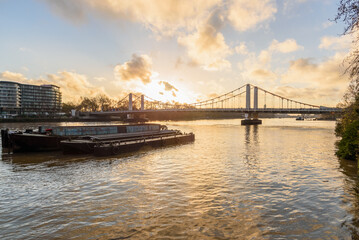 Fototapeta premium Suspension road bridge over a river at sunset in winter. Anchored barges are in foreground. London, UK.