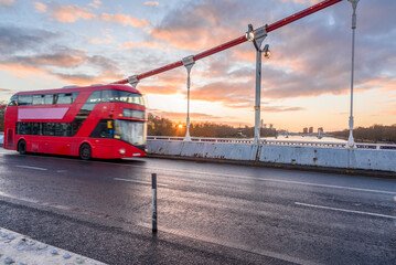 Red double decker bus transiting over a suspension bridge at sunset in winter. A riverside park is visible in background. Chelsea bridge, London, UK.