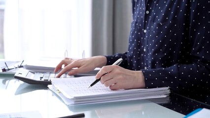 Businesswoman making notes while reviewing ring folder of financial documents with magnifying glass...