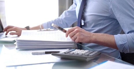 Fototapeta premium A businessman is using a calculator and a magnifying glass to analyze financial documents at his glass desk in a modern and fair office setting, focusing on aspects of audit and taxes in business