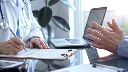 Doctor woman wearing white medical coat is consulting patient in clinic office while both are sitting at the glass desk. Physician is listening and making notes. Medicine concept