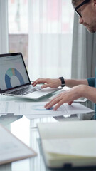 Fototapeta premium Businessman wearing green shirt and glasses is working with laptop computer and data analysis charts. Vertical business portrait