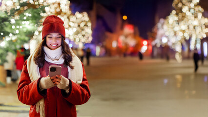 Young woman using smartphone while walking in the evening on Xmas Eve, enjoying festive vibe in the city and Christmas time