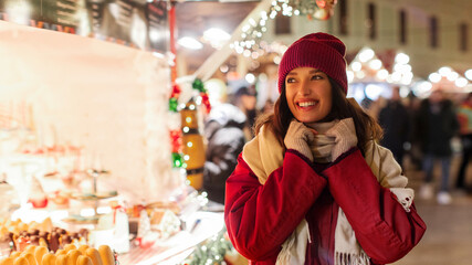 Obraz premium Portrait of happy lady walking at Christmas market in the evening, looking at various candies and gingerbread sweets, enjoying festive atmosphere