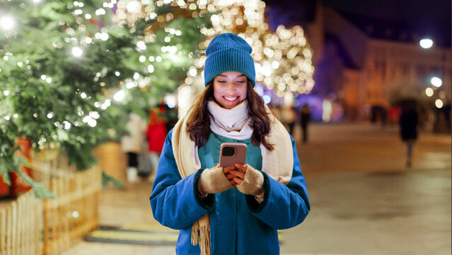 Young woman using smartphone while walking in the evening on Xmas Eve, enjoying festive vibe in the city and Christmas time