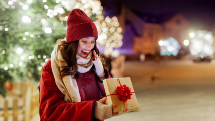Overjoyed young European woman rejoicing opening gift box, standing in front of decorated Christmas tree with garland lights outdoors, free space