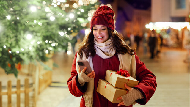 Modern holiday shopping. Happy lady holding gift boxes and using smartphone while standing outdoors near Xmas tree and festive decorations