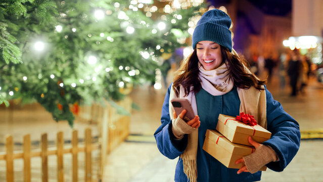 Modern holiday shopping. Happy lady holding gift boxes and using smartphone while standing outdoors near Xmas tree and festive decorations