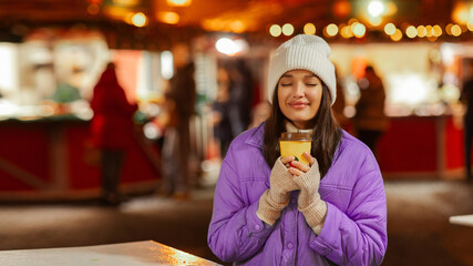 Happy lady enjoying drinking takeaway cup of mulled wine or hot tea outdoors on street with festive illumination, walking on Christmas Eve
