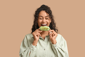 African-American woman eating tasty eclair on beige background