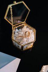 Two gold wedding rings in a glass box filled with decorative pearls on black background