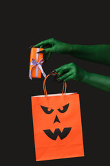 Green hands of witch with gift box and shopping bag on black background. Halloween celebration