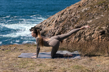 Woman practicing yoga on a mat by the ocean, stretching in a serene environment with rocky cliffs and gentle waves, embodying tranquility and mindfulness