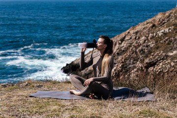 Woman practicing meditation and stretching on a yoga mat by the ocean, enjoying a refreshing drink, surrounded by nature and scenic coastal views, promoting wellness and relaxation