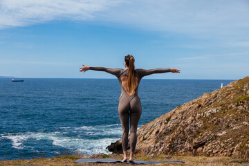Woman practicing yoga on a mat by the ocean, stretching arms wide open, embracing nature and tranquility, with waves crashing against the rocky shore and a clear blue sky