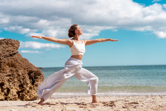 Young woman practicing Warrior Pose yoga on sandy beach with arms outstretched stretching and focusing on balance surrounded by peaceful ocean and cloudy sky - Powered by Adobe