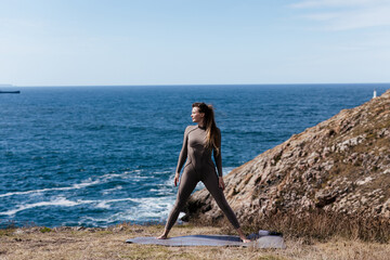 Woman practicing yoga on a mat by the ocean, showcasing a strong pose against a backdrop of rocky cliffs and serene water, embodying tranquility and strength in nature
