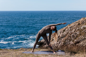 Woman practicing yoga outdoors, stretching gracefully on a mat, with a stunning ocean view and rocky landscape, embodying tranquility and connection to nature