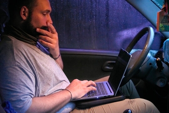 Hispanic male traveler using a laptop sitting in his car, it is nighttime and raining outside. Concepts: digital nomad, remote work and working while traveling, freedom lifestyle.