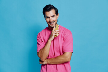 Smiling man wearing a pink t-shirt stands confidently against a blue background, holding a phone near his face. Engaging and cheerful studio portrait image.