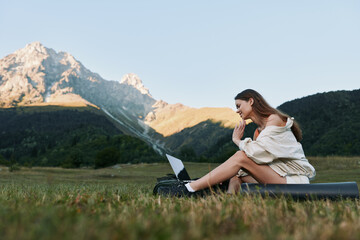 A casual outdoor workspace scene shows a woman with a laptop in a meadow under mountains. She smiles softly, focused on her screen, creating a calm, productive atmosphere.