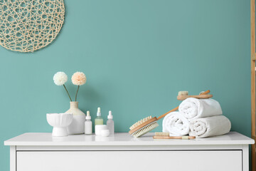 Different massage brushes with folded towels, cosmetic products and flowers on commode near blue wall in bathroom